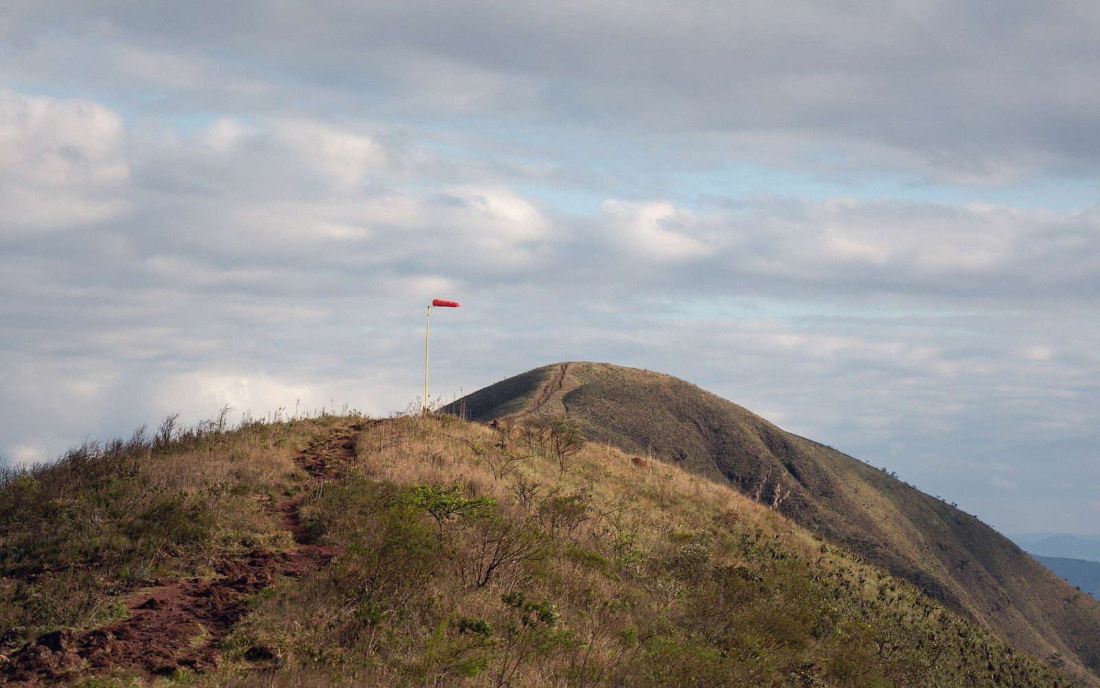 A red windsock on a pole marks a grassy mountain ridge under cloudy skies